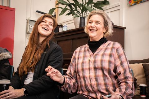 Two women laughing together on the couch, an upright piano behind them